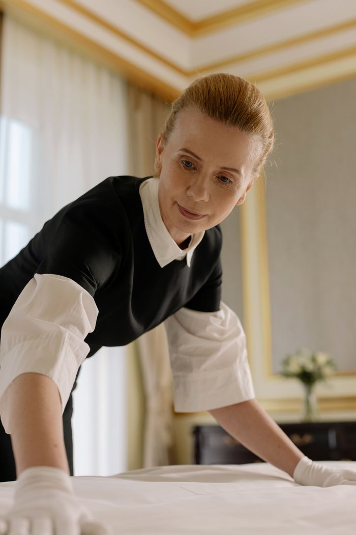 services-04 A housekeeper in uniform skillfully arranges bed linen in a luxurious hotel room.