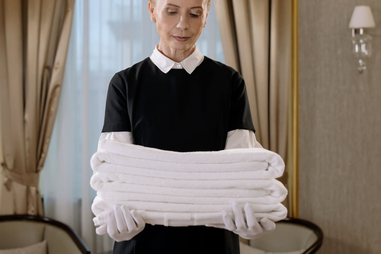 hero-img Professional housekeeper holding a stack of fresh towels in a luxurious hotel room, showcasing elegance and service.